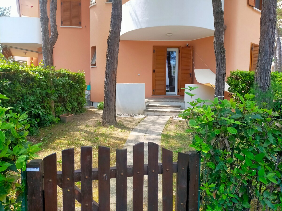 A wooden gate opens to a pathway leading to the entrance of a two-story building, flanked by lush greenery. The exterior features soft peach-colored walls and shuttered windows. A covered porch area is visible, providing a welcoming entry into the residence.