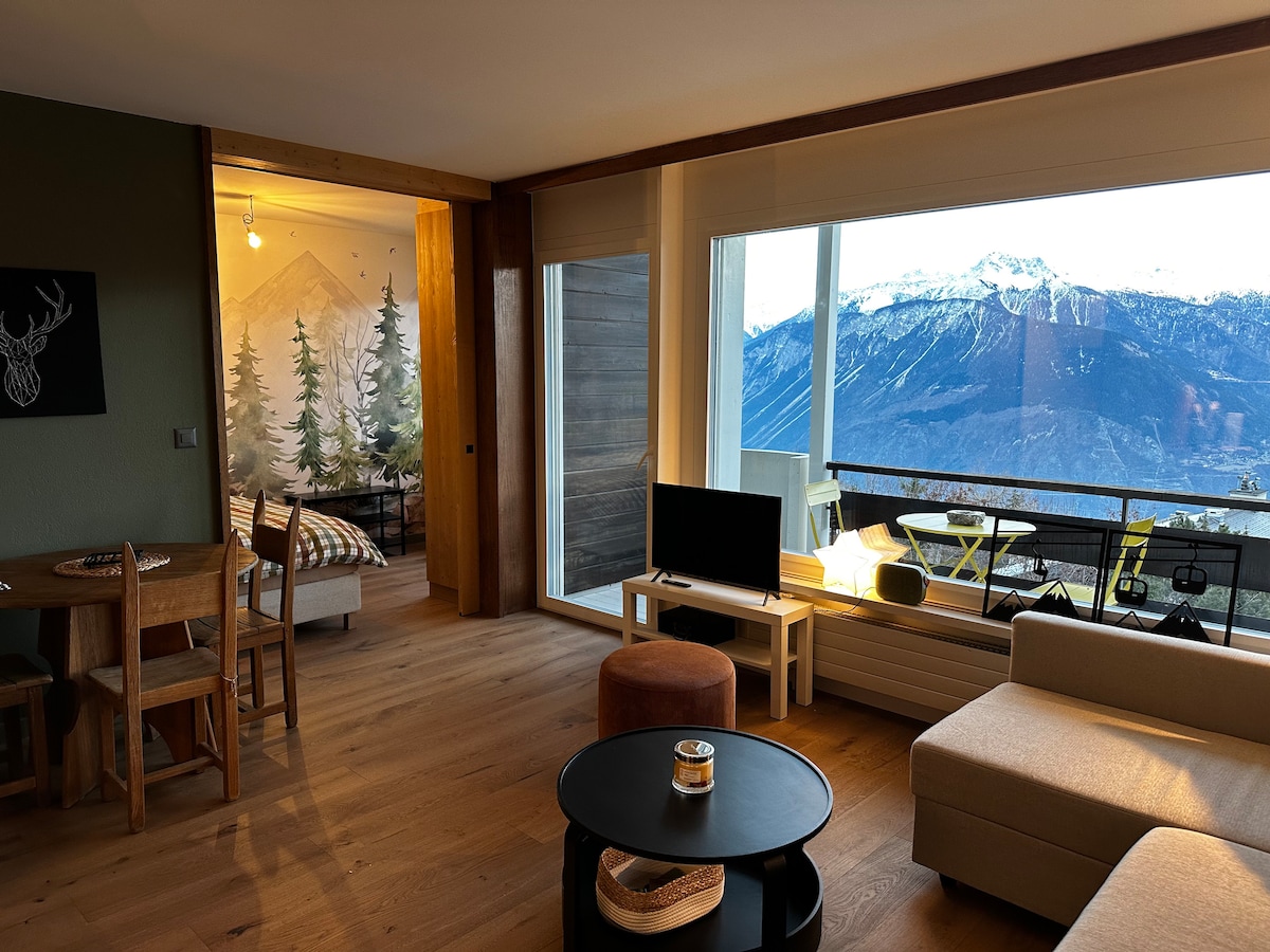 An open space features large windows that offer views of the mountains. A comfortable seating area is complemented by a dark circular table. The adjacent bedroom's wall showcases a nature-themed mural, creating a harmonious connection with the outdoor scenery.