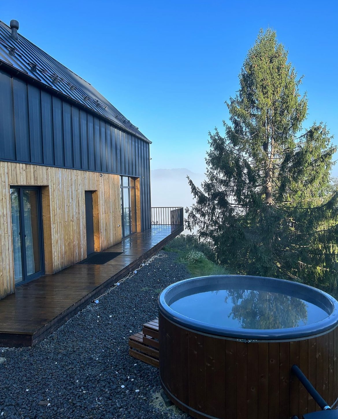 A wooden deck extends from the cottage, showcasing a circular hot tub adjacent to a lush green tree. The structure features a mix of wooden and metal exterior, with large glass doors providing views of the surrounding mountains and an expansive blue sky.