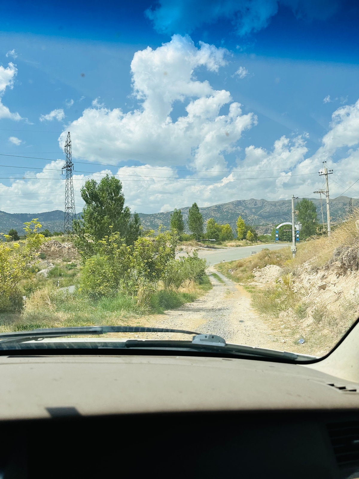 A scenic country road is visible from the perspective of a vehicle, flanked by greenery and rolling hills. A blue sky dotted with clouds creates a serene atmosphere. Power lines cross the landscape, leading towards distant mountains in the background.