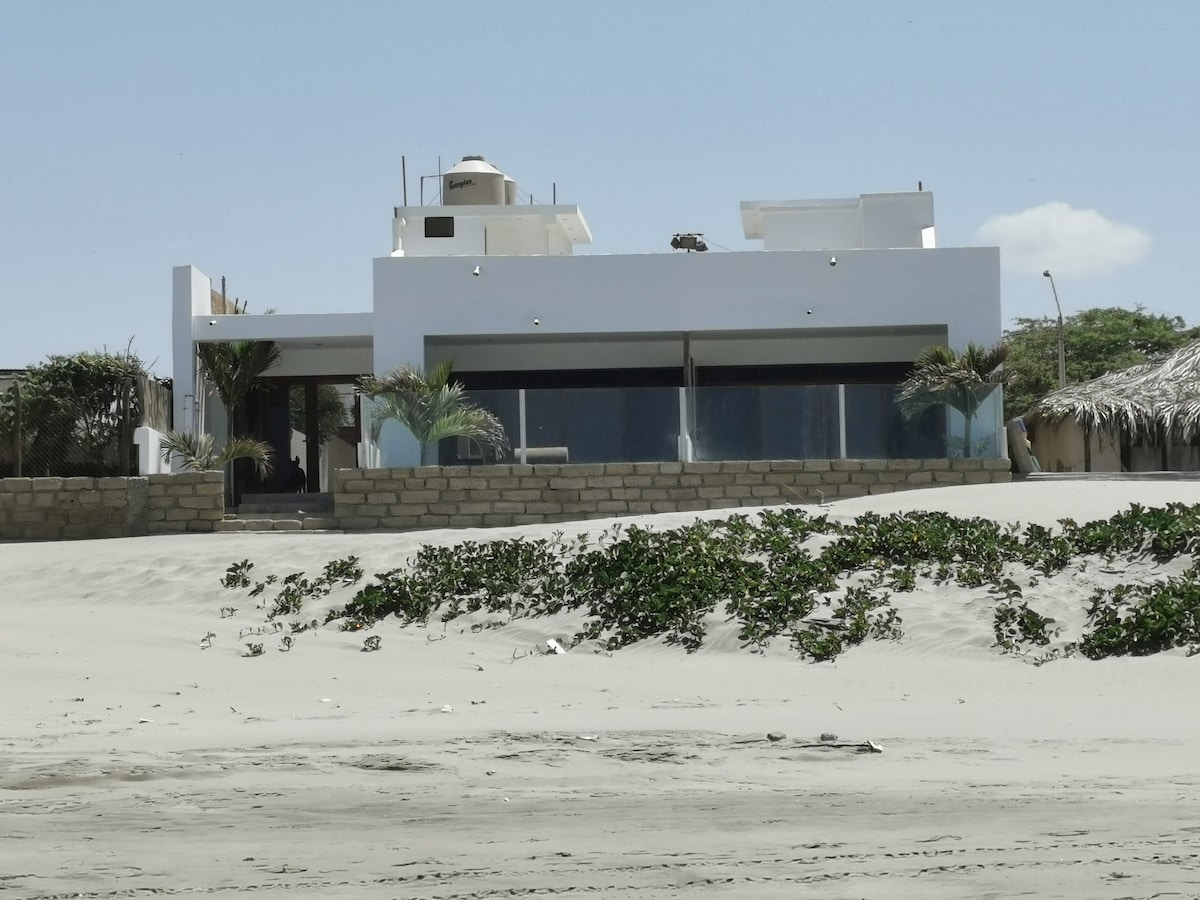 A modern beachfront house is displayed, featuring expansive glass windows that provide views of the surrounding landscape. The exterior includes a smooth white facade, complemented by palm trees and a sandy area leading up to the property.