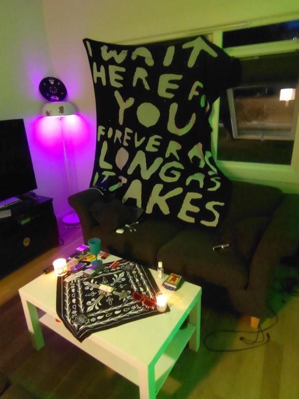A cozy seating area features a dark sofa with a large blanket draped over it, displaying bold text. A white coffee table is decorated with art supplies and candles, while ambient lighting casts a soft glow in the room.
