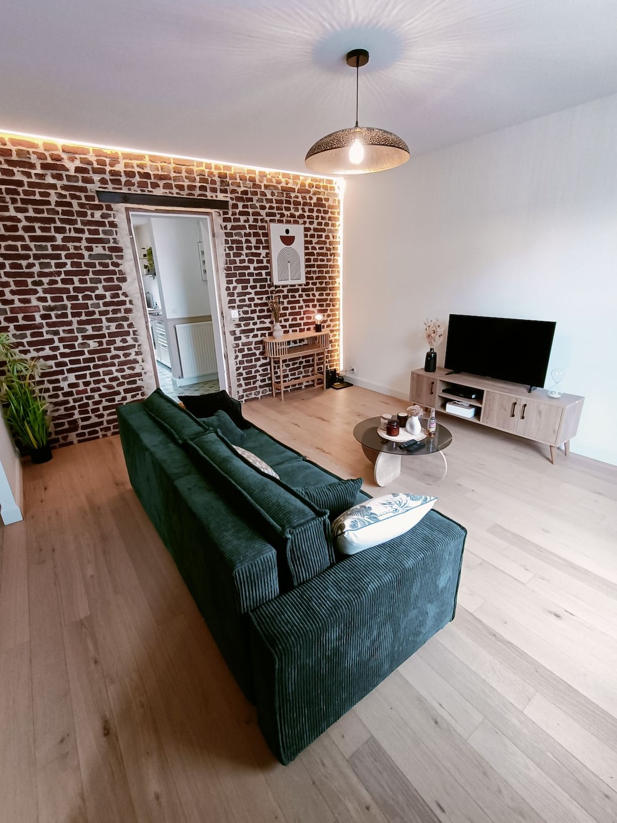 A modern living area features a deep green sofa facing a low coffee table and a flat-screen television. Exposed brick walls add texture, and natural light enhances the open layout. A small dining area with chairs is visible in the background, offering additional seating.