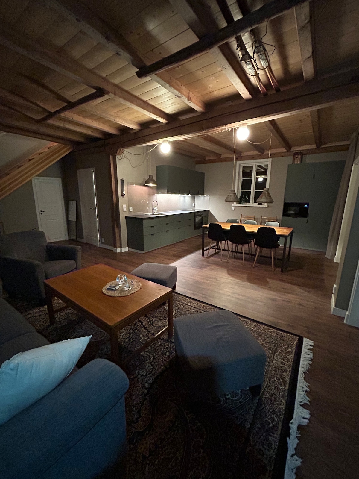 An open living space features a cozy seating area with a sofa and armchair facing a wooden coffee table. A dining table for four is situated nearby, with modern kitchen cabinets visible in the background. Soft lighting highlights the wooden ceiling and warm flooring.