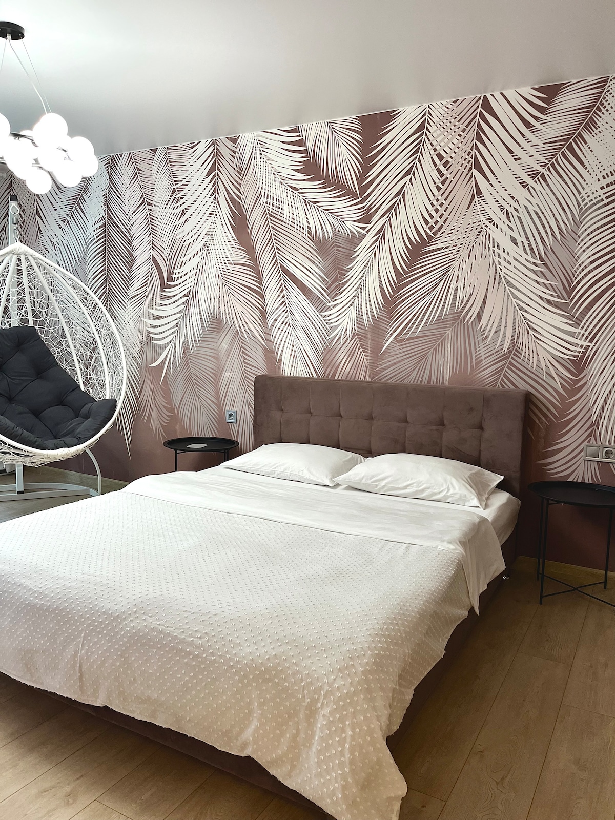 A spacious bed with a soft white bedding set is situated against a decorative wall featuring a leaf pattern in muted tones. A hanging swing chair is positioned nearby, creating a relaxed atmosphere. Light-colored wooden flooring complements the overall design.