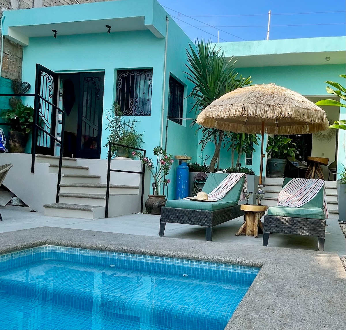 A tranquil outdoor space features a small pool surrounded by lounge chairs, each with a patterned cushion. A thatched umbrella provides shade nearby, while vibrant greenery and potted plants add a natural touch to the setting.