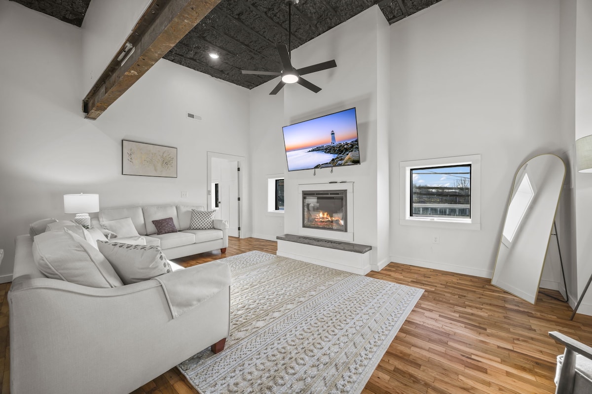 A spacious living area is highlighted by a modern fireplace and television above a sleek mantel. Light-colored sofas are oriented towards the fireplace, with a large area rug adding warmth to the hardwood floor. Bright windows provide ample natural light.