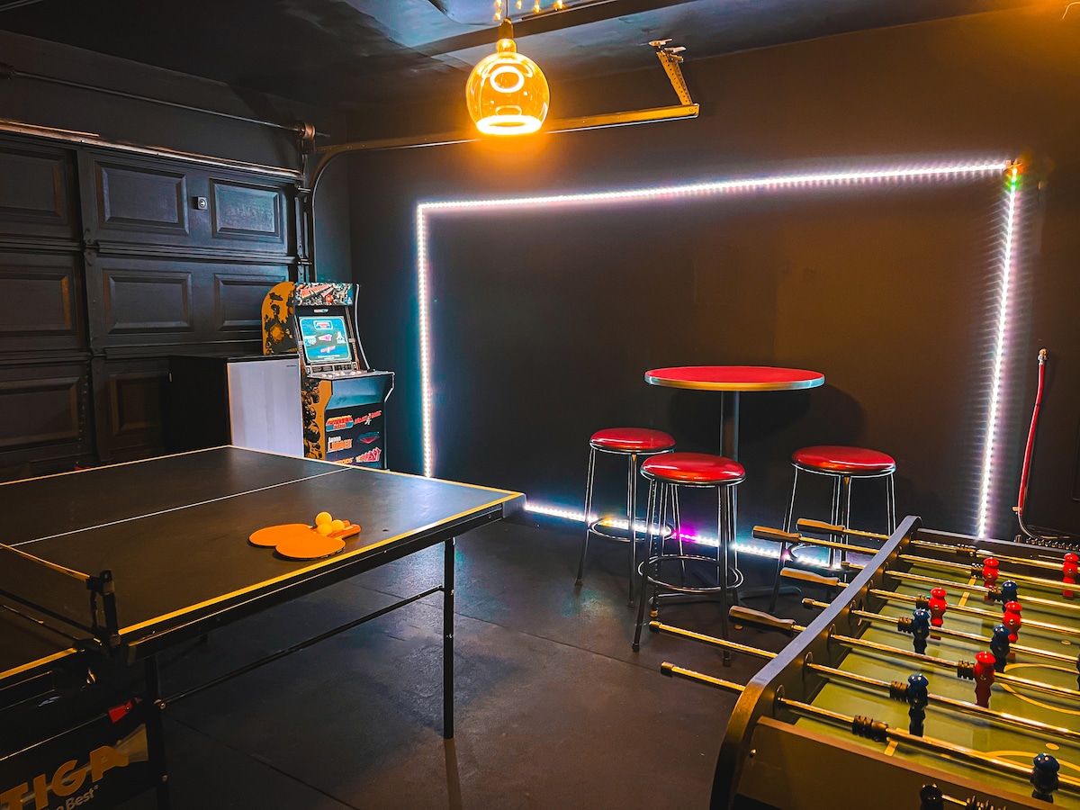 The game room is equipped with a full-size ping pong table and a foosball table. A bar table with red stools provides seating, while a classic arcade game adds entertainment. Color changing LED lights illuminate the area, creating a vibrant environment for leisure activities.