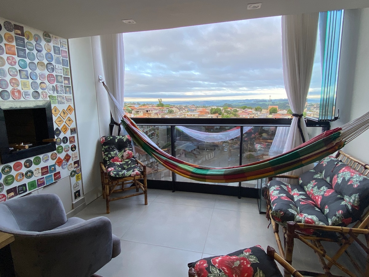 A comfortable seating area is presented with a hammock and floral-patterned chairs, offering a cozy space. Large glass windows provide a scenic view of the surrounding area, while light curtains allow for natural light to filter in.