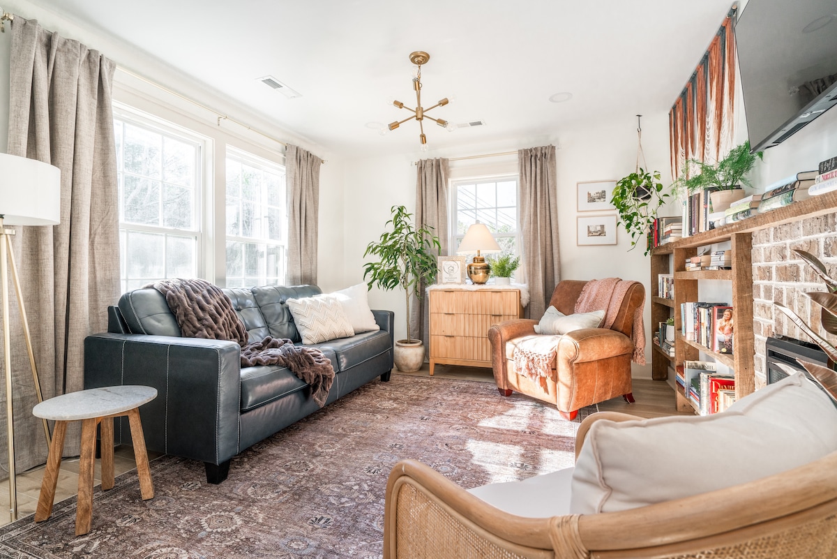 A living room features a leather sofa and two accent chairs, complemented by a wooden side table. A large window allows natural light to illuminate a cozy rug. Shelves filled with books and greenery create a serene atmosphere, accompanied by a stylish light fixture overhead.