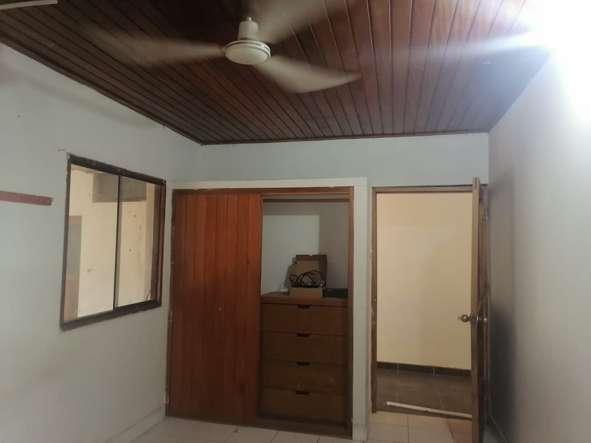 A simple room is depicted, featuring a wooden ceiling and a ceiling fan. A wooden closet with drawers is visible, along with an open door leading to another space. Natural light enters through a window on the opposite wall.