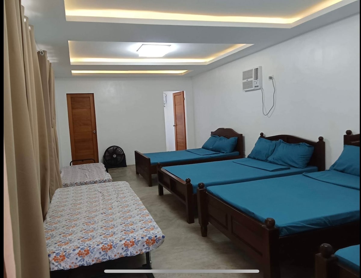 A well-lit accommodation space features multiple beds with blue bedding and wooden frames. Light-colored walls enhance the airy feel. A large window covered by beige curtains allows natural light to enter, while a fan is positioned nearby for comfort. A door leads to another area.