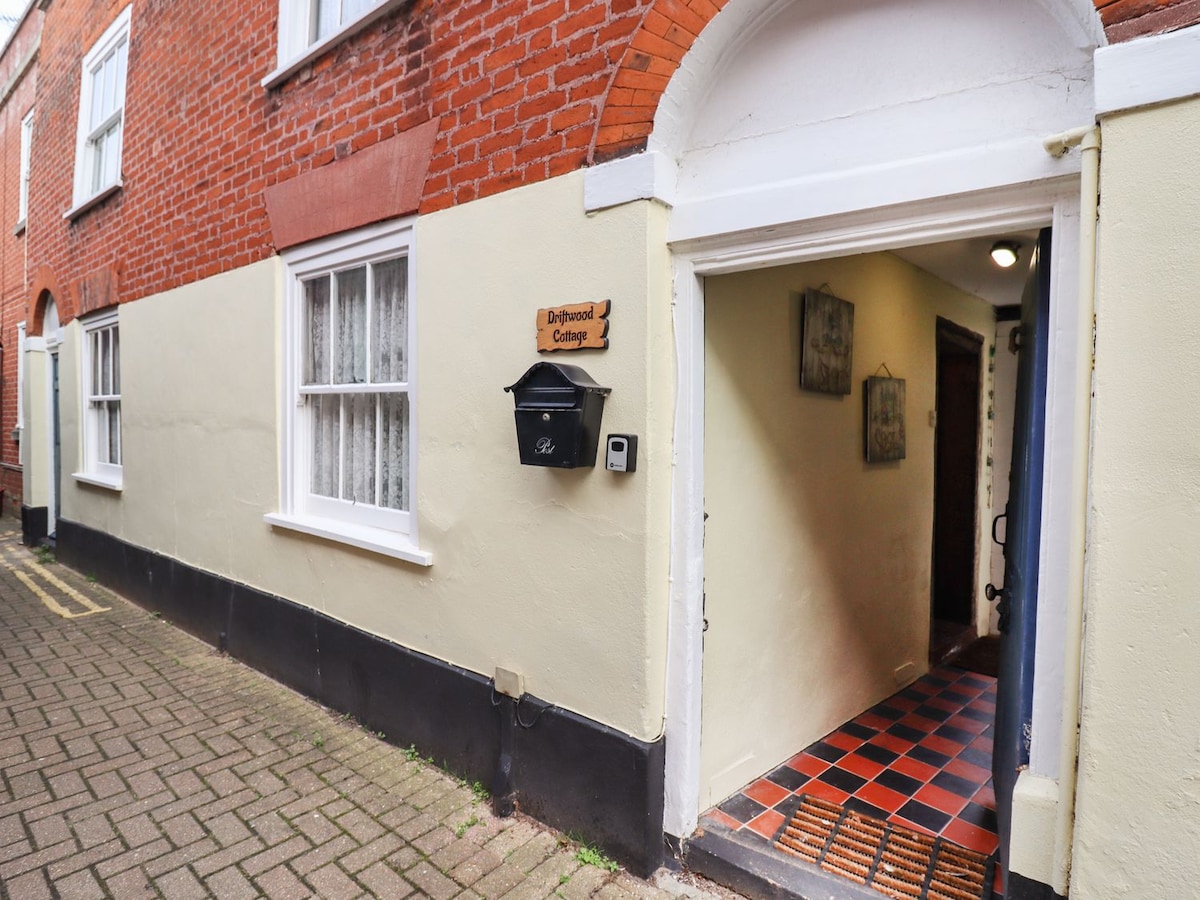 The entrance to the cottage features a covered area with a distinctive archway. A black mailbox is mounted on the wall, displaying the name of the cottage. The pathway is lined with textured red and black tiles, leading into the interior.