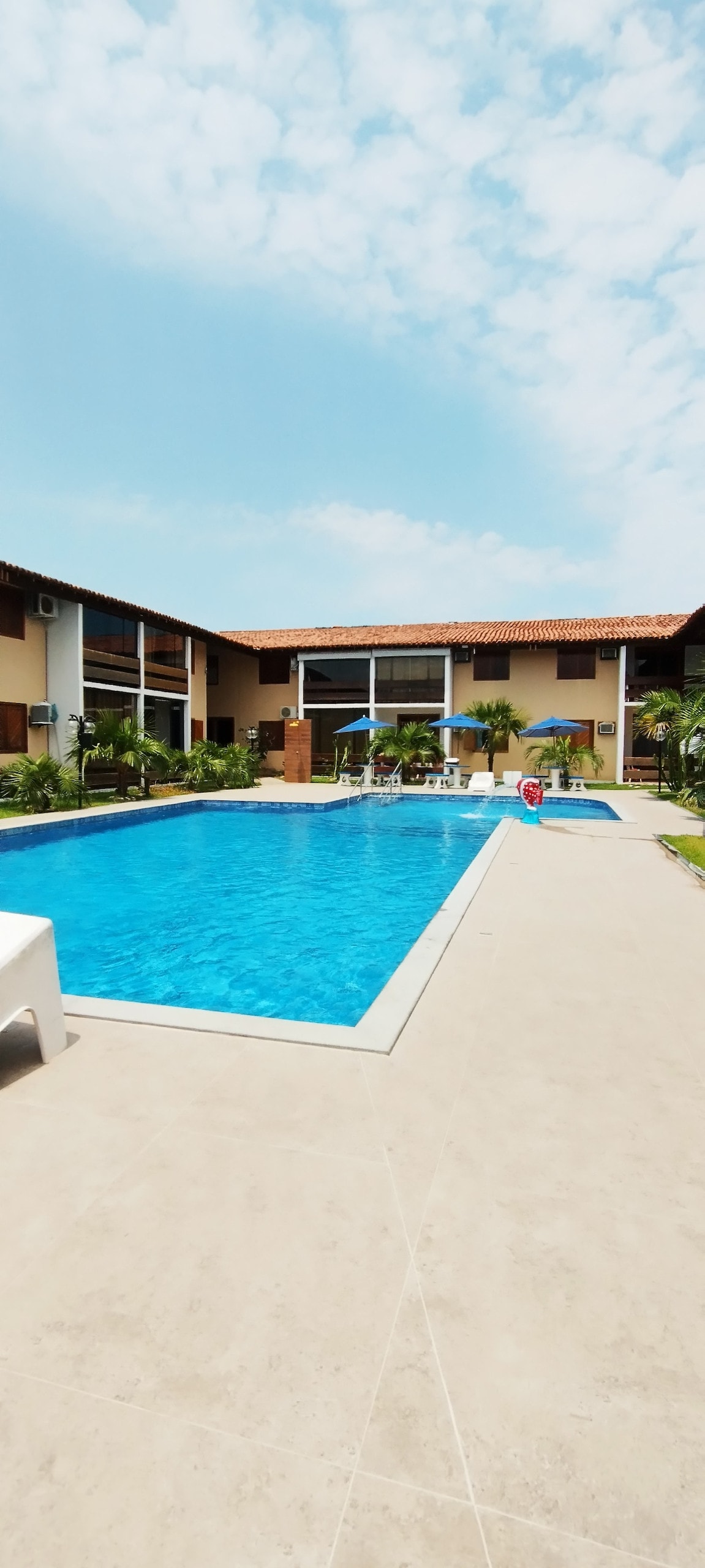 A spacious outdoor area features a large swimming pool surrounded by palm trees and lounge chairs. The clear blue water contrasts with the light beige tiles. The building's exterior showcases multiple levels with large windows, highlighting the welcoming atmosphere of the shared space.