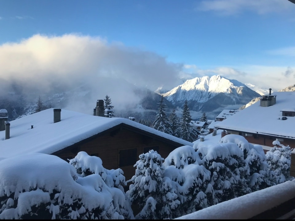 A scenic winter landscape is visible, featuring snow-covered rooftops and trees. Majestic mountains rise in the background, partially obscured by soft clouds. The tranquil atmosphere is highlighted by the white snow blanketing the surroundings, creating a peaceful winter scene.