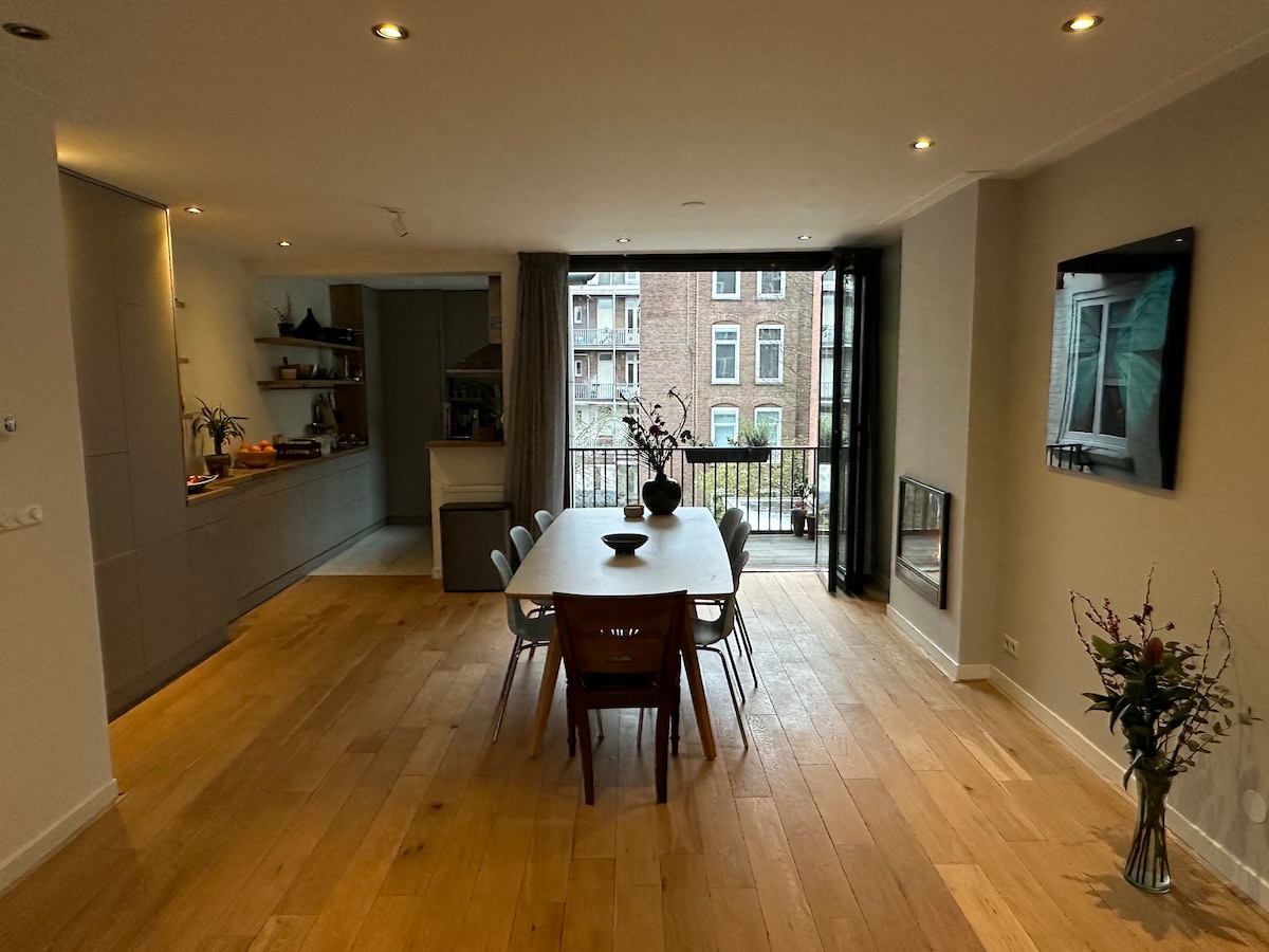 A dining area showcases a long wooden table surrounded by six chairs. Natural light from large glass doors illuminates the space, which features wooden flooring and a minimalist kitchen in the background. Decorative elements include a floral arrangement on the table.