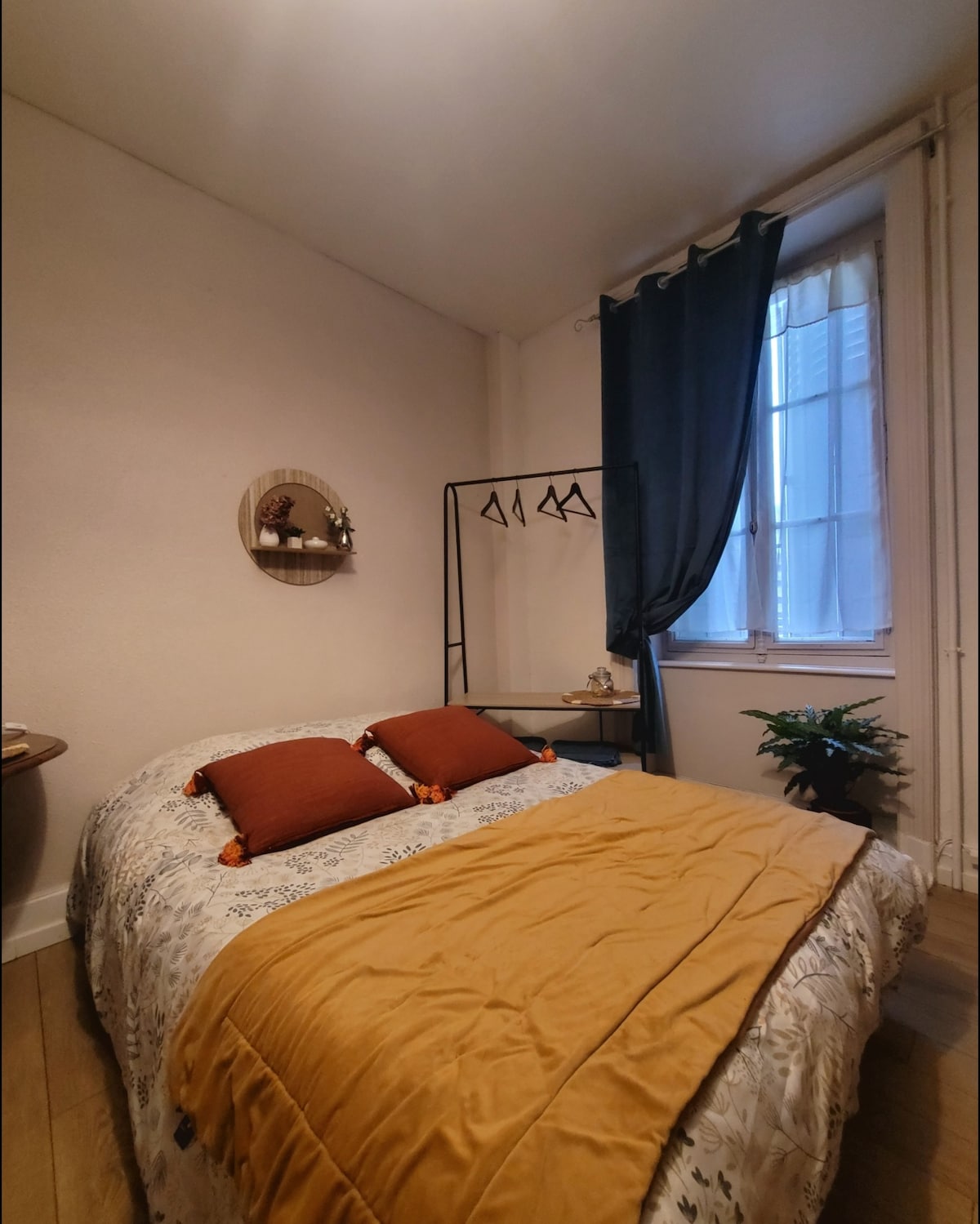A comfortable sleeping area features a spacious bed adorned with colorful pillows and a textured blanket. A minimalist clothing rack stands alongside a small table, while a plant is positioned in a corner. Natural light enters through a window, partially covered by sheer curtains.
