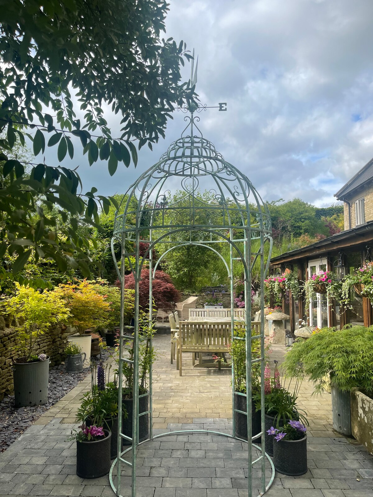 A charming garden space is highlighted by a decorative metal gazebo, surrounded by lush greenery and a variety of flowering plants. A wooden dining table and chairs can be seen in the background, nestled under the shade of nearby trees.