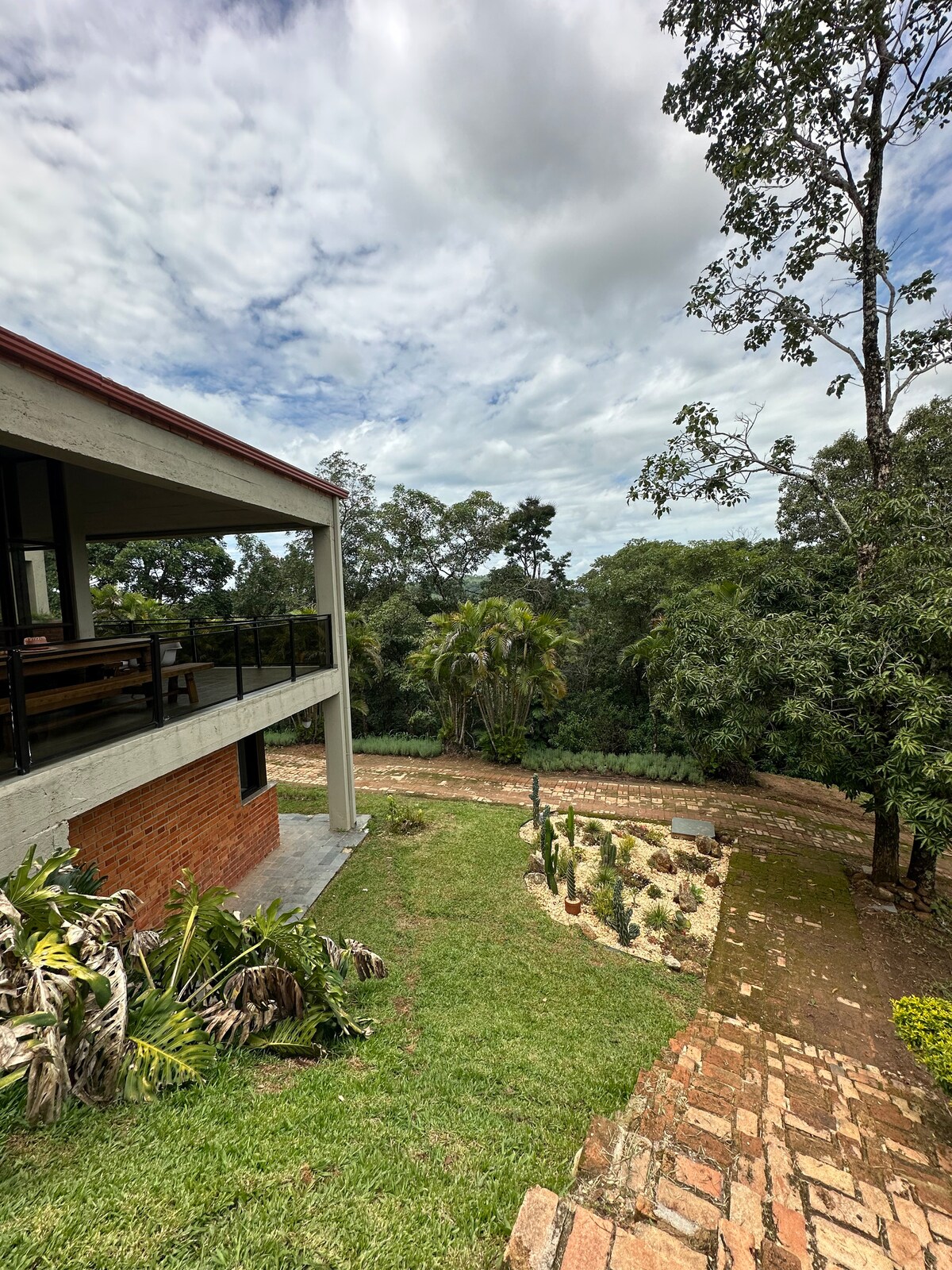 A modern house is seen on the left with large windows and a balcony, overlooking a lush garden filled with greenery. A stone path winds through the yard, leading towards a densely wooded area in the background under a partly cloudy sky.