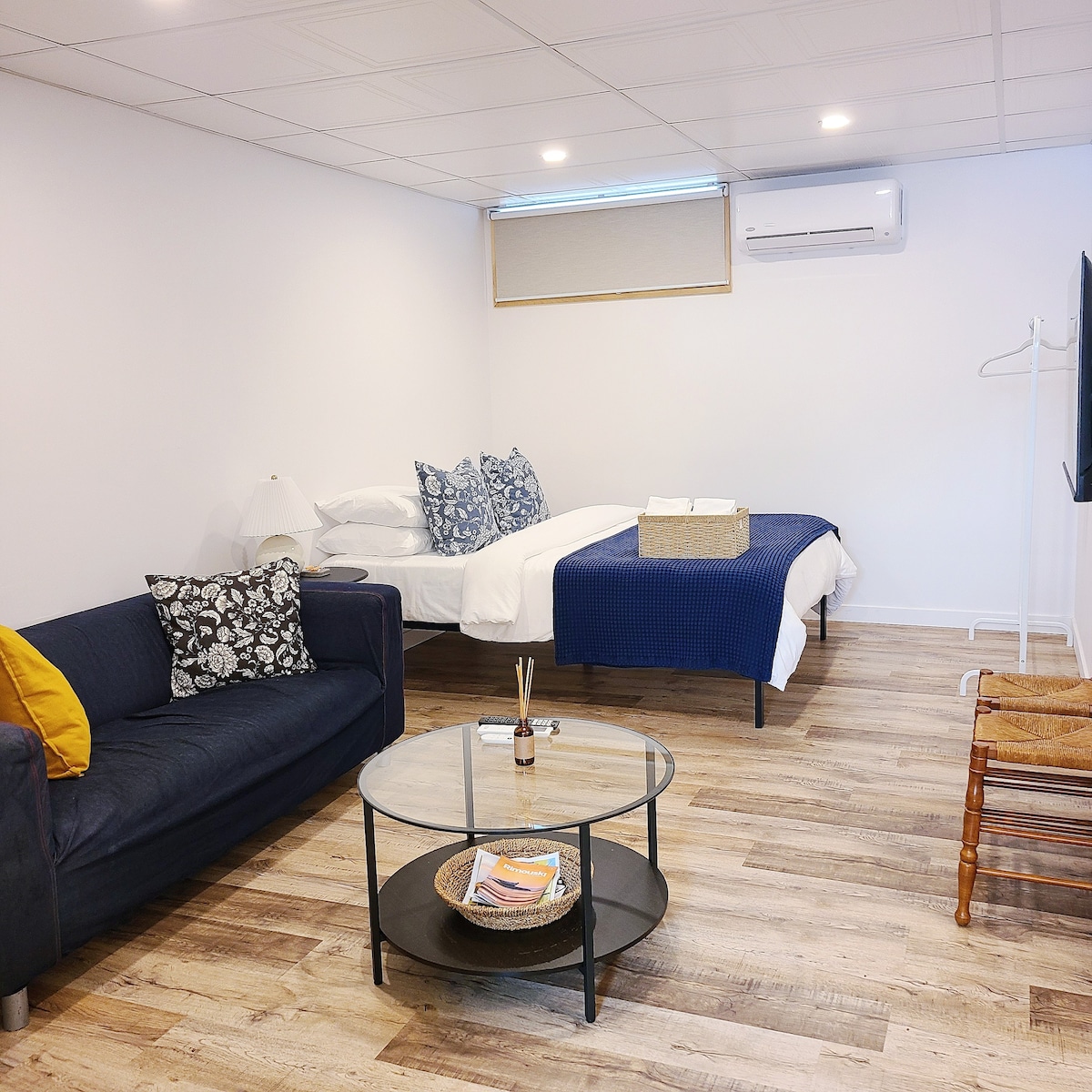 A comfortable studio space features a spacious bed adorned with decorative pillows. A dark sofa sits near a round coffee table, and a TV is positioned against the wall. Natural light enters through a small window, enhancing the cozy atmosphere.