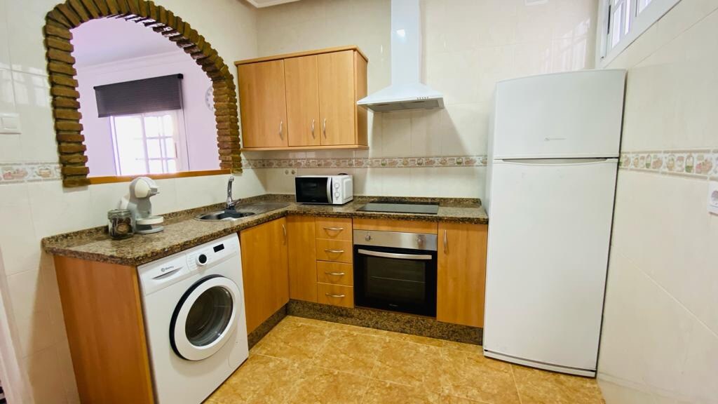 The kitchen features a modern design with wooden cabinets and granite countertops. Essential appliances are included, such as a refrigerator, microwave, oven, and washing machine. Natural light enters through a window, illuminating the space and creating a functional area for meal preparation.