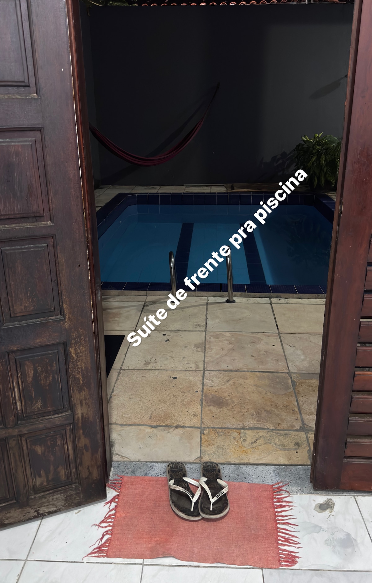 An inviting view of the private suite entrance is framed by a pair of wooden doors. A small rug lies on the tiled floor, leading to a tranquil pool area, where a hammock is hung beside the water, surrounded by lush greenery.