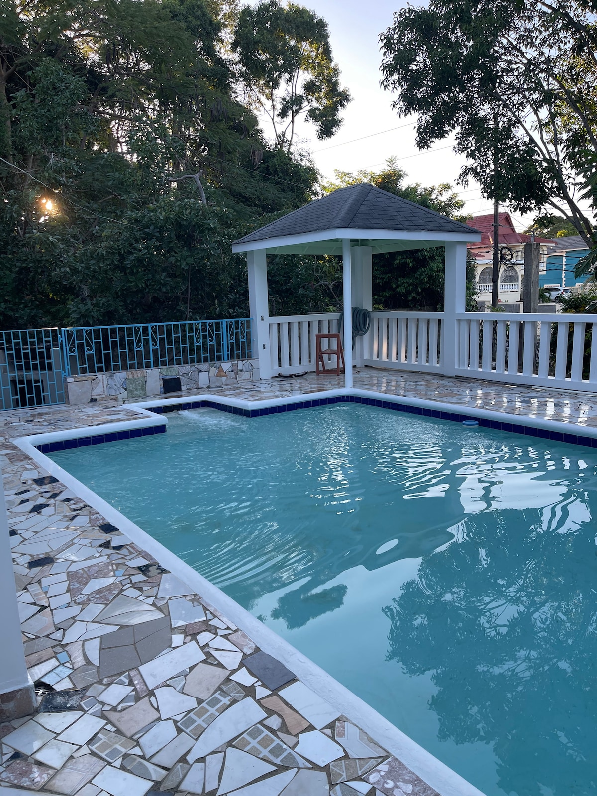 A private swimming pool is framed by a stone patio, showcasing intricate tile patterns. A gazebo with a sloped roof offers shaded seating nearby. Lush greenery surrounds the area, creating a serene atmosphere, with morning light softly filtering through the trees.