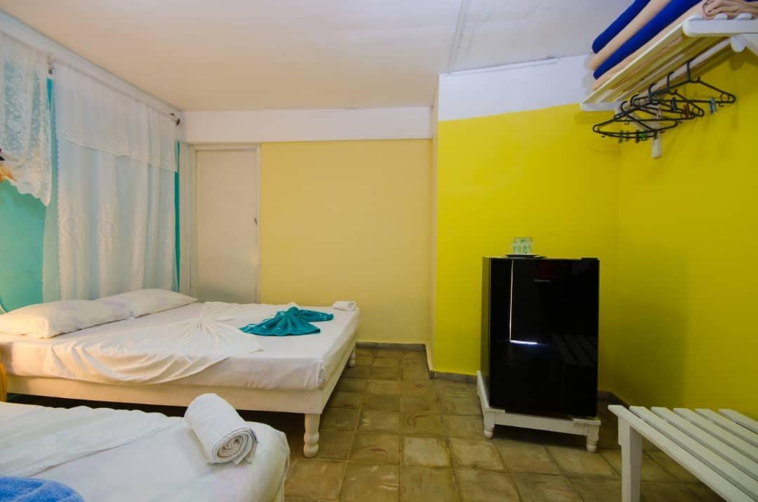 A simple sleeping area features two spacious beds with light bedding. A small black refrigerator is placed against the wall, while a white bench is positioned nearby. The walls are painted in soft yellow and teal, creating a bright and cheerful environment.