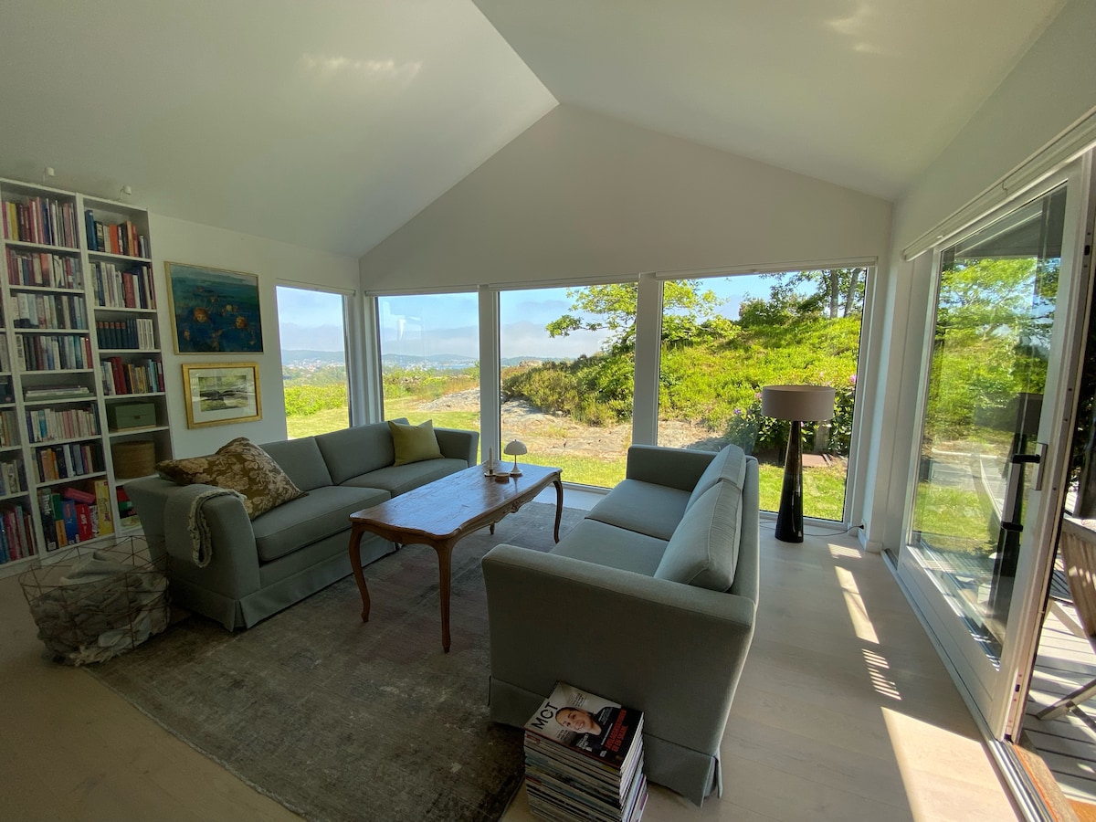 A spacious living area features large windows allowing natural light to fill the room and offering views of the surrounding landscape. Comfortable seating is arranged around a central wooden coffee table on a soft rug, complemented by a standalone lamp and a bookshelf with an assortment of books.