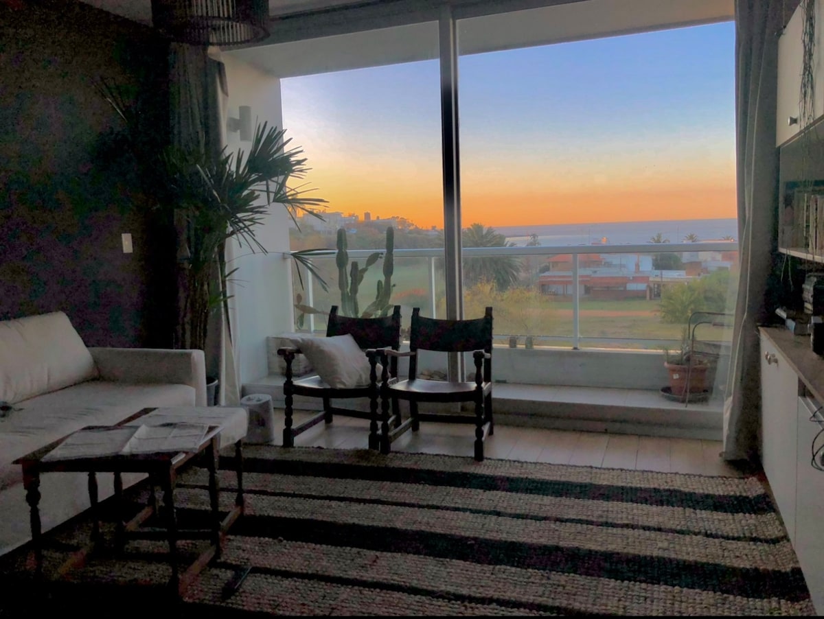 A living area features light-colored furniture arranged around a dark woven rug. Floor-to-ceiling windows provide a view of the sunset over the ocean, enhancing the space's tranquility. Potted plants and a couple of wooden chairs add to the relaxed atmosphere.