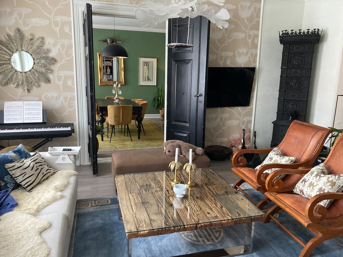 A spacious living area is presented, featuring a wooden coffee table at the center. Comfortable seating options include two leather armchairs and a cozy sofa. A piano and a large mirror are visible, with a dining space in the background highlighted by a green wall and ornate decor.