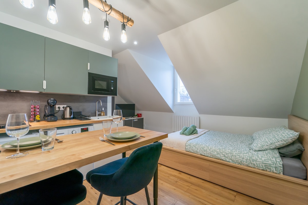 A studio space features a compact kitchenette with modern appliances, including a microwave and coffee maker. A dining table is set for two, accompanied by blue chairs. A sleeping area showcases a neatly made bed with fresh linens, complemented by natural light from the window.