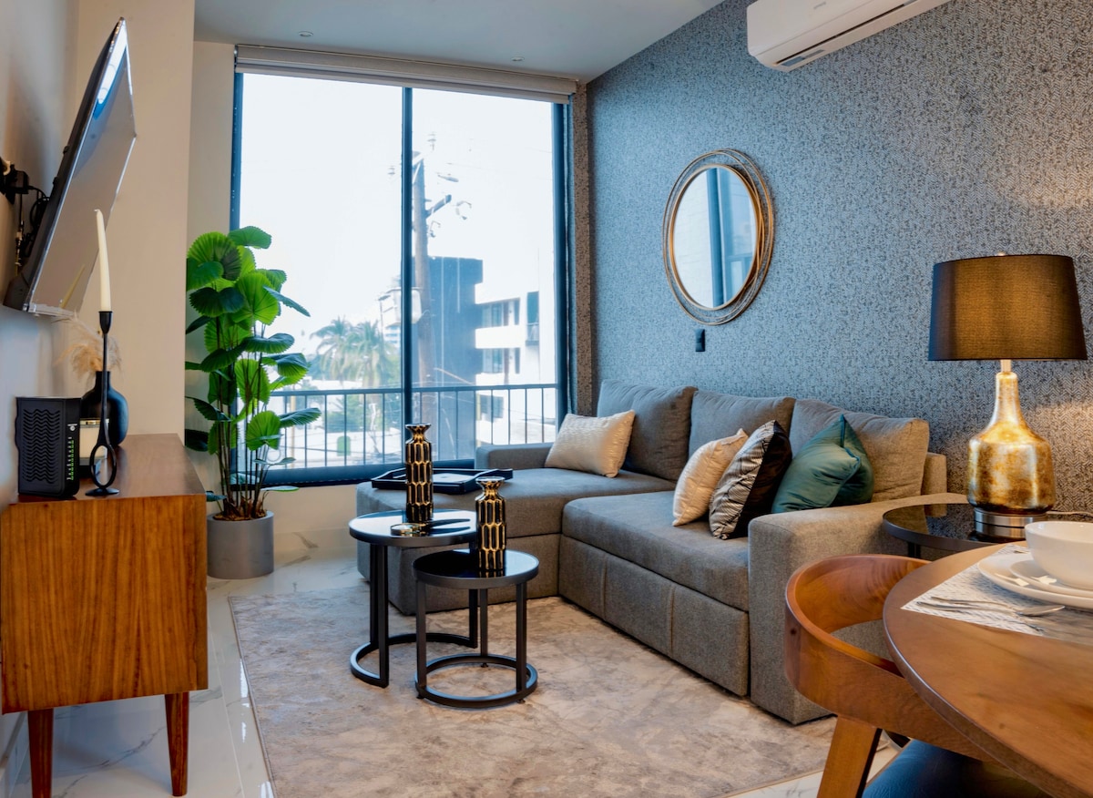 A modern living area features a comfortable gray sectional sofa adorned with decorative pillows. Two round side tables are placed alongside the sofa, and a large window allows natural light to fill the space, revealing a glimpse of the outdoor scenery. A dining area is visible in the background.