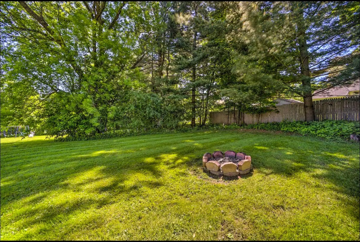 A grassy outdoor area is surrounded by mature trees, providing a serene backdrop. A circular fire pit made of stones is situated on the lawn, creating an inviting space for gatherings. Sunlight filters through the trees, casting gentle shadows on the ground.