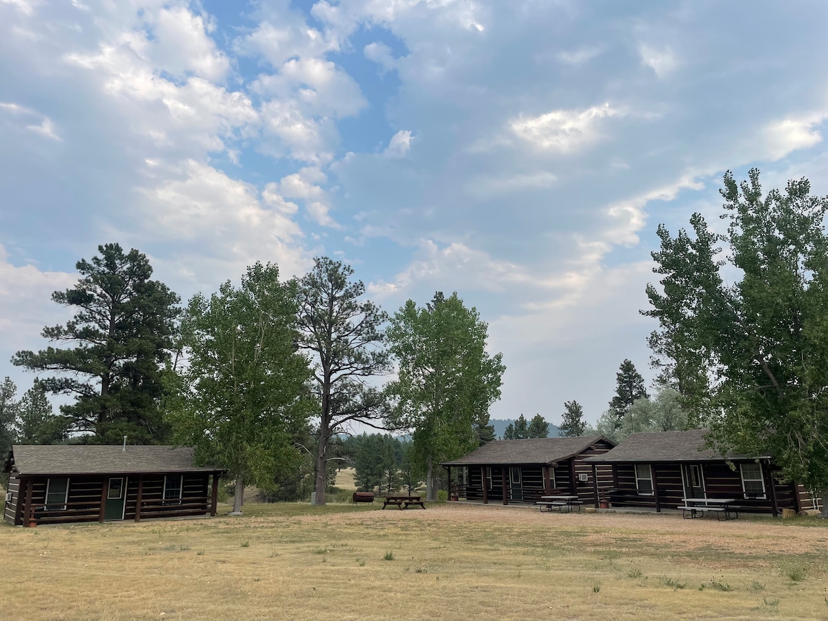 A serene landscape features three rustic log cabins set amidst green trees and grassy areas. The sky is partly cloudy, creating a calm atmosphere. A picnic table is positioned on the grassy knoll, providing an inviting spot for outdoor gatherings.