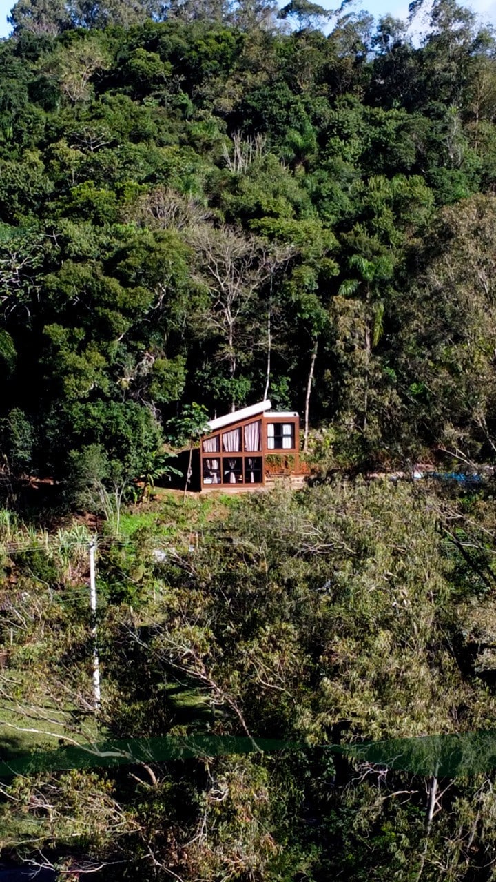 A wooden cabin is nestled within lush greenery, surrounded by dense trees. Large glass windows reflect the natural landscape, while a gently sloping terrain leads to the structure, emphasizing its integration with the outdoor environment.