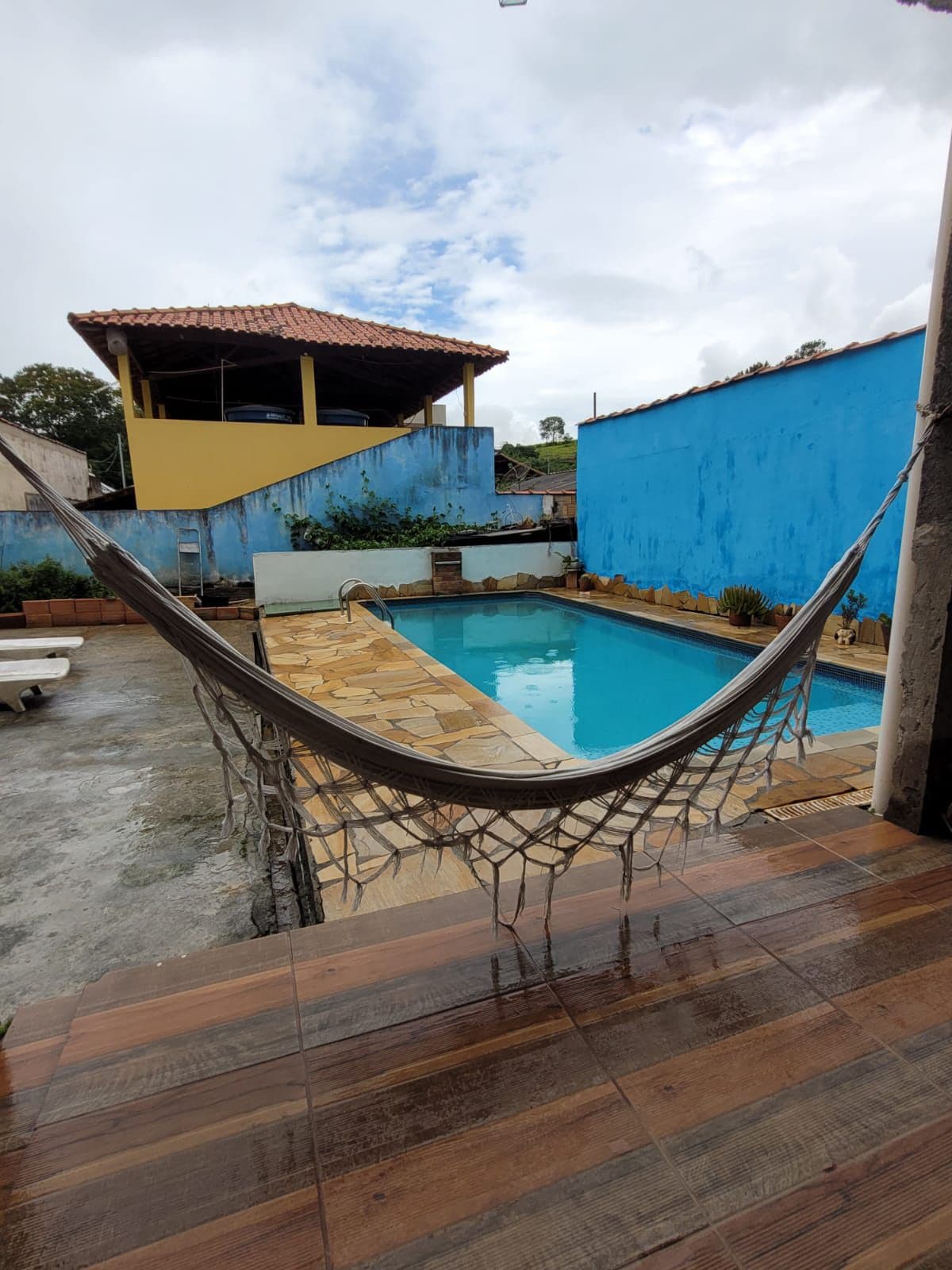 A serene outdoor space features a clear swimming pool surrounded by tiled areas and potted plants. A hammock is suspended nearby, inviting relaxation. The walls are painted in vibrant blue, with a two-story structure visible in the background.
