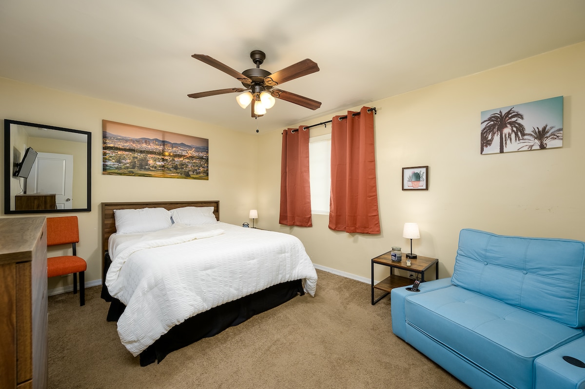 The master bedroom features a spacious bed with a white quilt and a cozy twin sleeper chair. A ceiling fan provides airflow, while decorative wall art enhances the ambiance. Two bedside lamps and a full-length mirror are included, creating a functional yet inviting space.