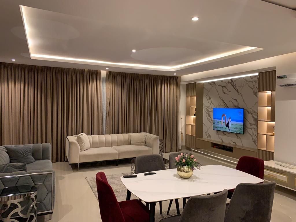 A spacious living area is illuminated by soft lighting, featuring a contemporary sectional sofa and an elegant dining table surrounded by upholstered chairs. A large television is mounted on a stylish feature wall, while rich curtains frame the windows, creating a refined atmosphere.