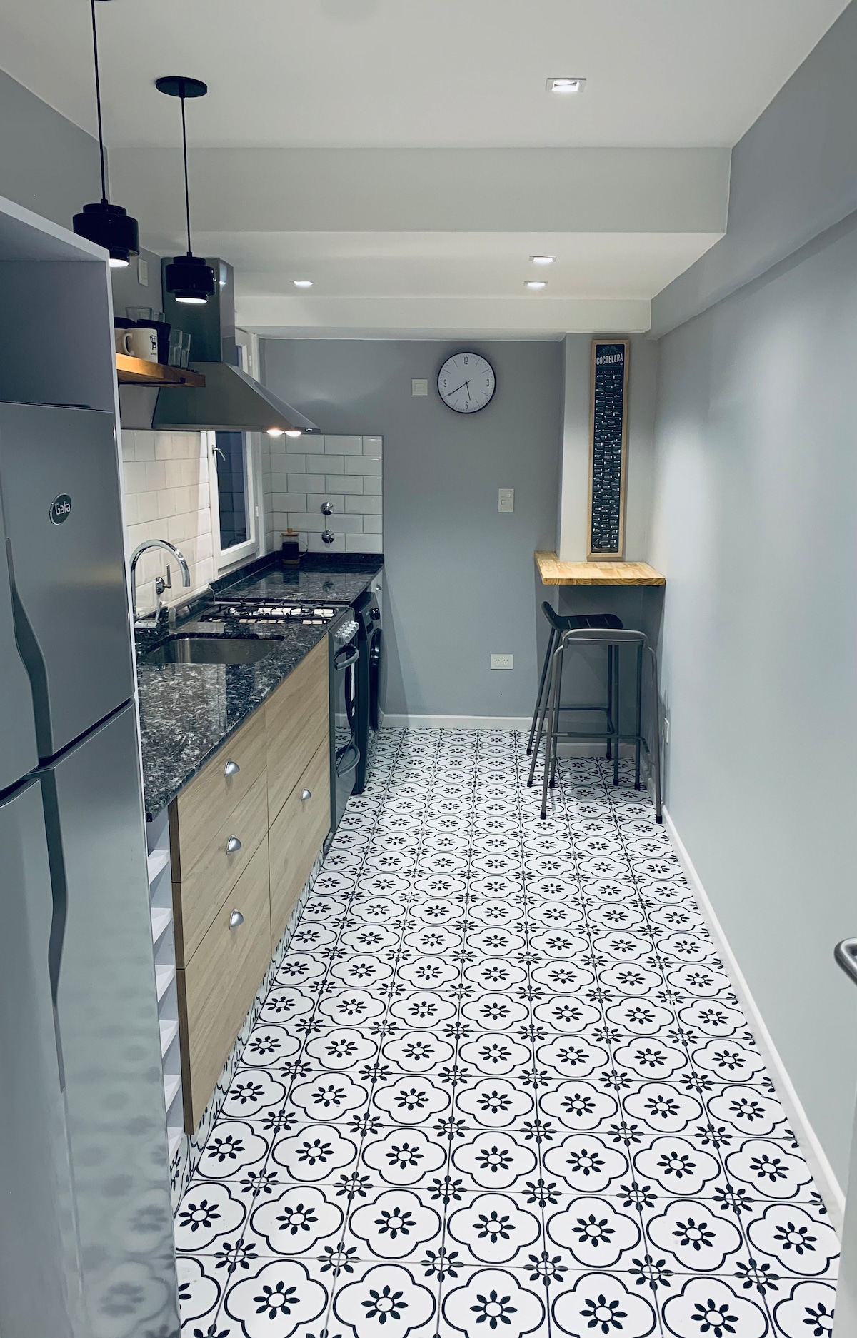 A modern kitchen features sleek cabinetry and a contrasting granite countertop. Stylish tiles with a black and white pattern line the floor. A small dining area with high stools is placed near the wall, and a clock is mounted above the countertop for convenience.