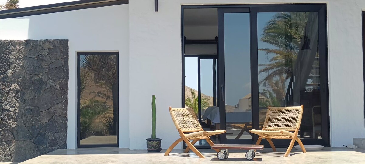 An outdoor seating area is featured at the entrance, showcasing two woven lounge chairs positioned facing large glass doors. A tall cactus sits in a decorative pot nearby, complemented by a smooth stone wall and a glimpse of palm trees beyond the entrance.