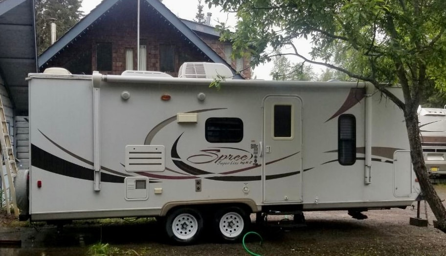 A travel trailer is positioned on a gravel surface, showcasing its sleek exterior with decorative graphics. Two axles are visible, along with a door on the side. Surrounding greenery and a nearby building are also present in the background.