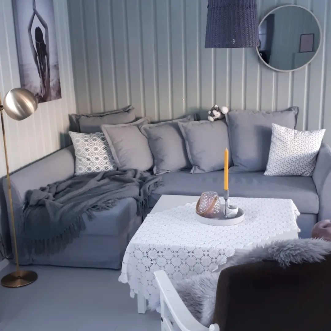 A cozy seating area is featured, with a gray sectional sofa adorned with various patterned cushions. A simple white table with a lace tablecloth is centered, holding a decorative tray with candles. A round mirror reflects light, enhancing the soft ambiance of the space.