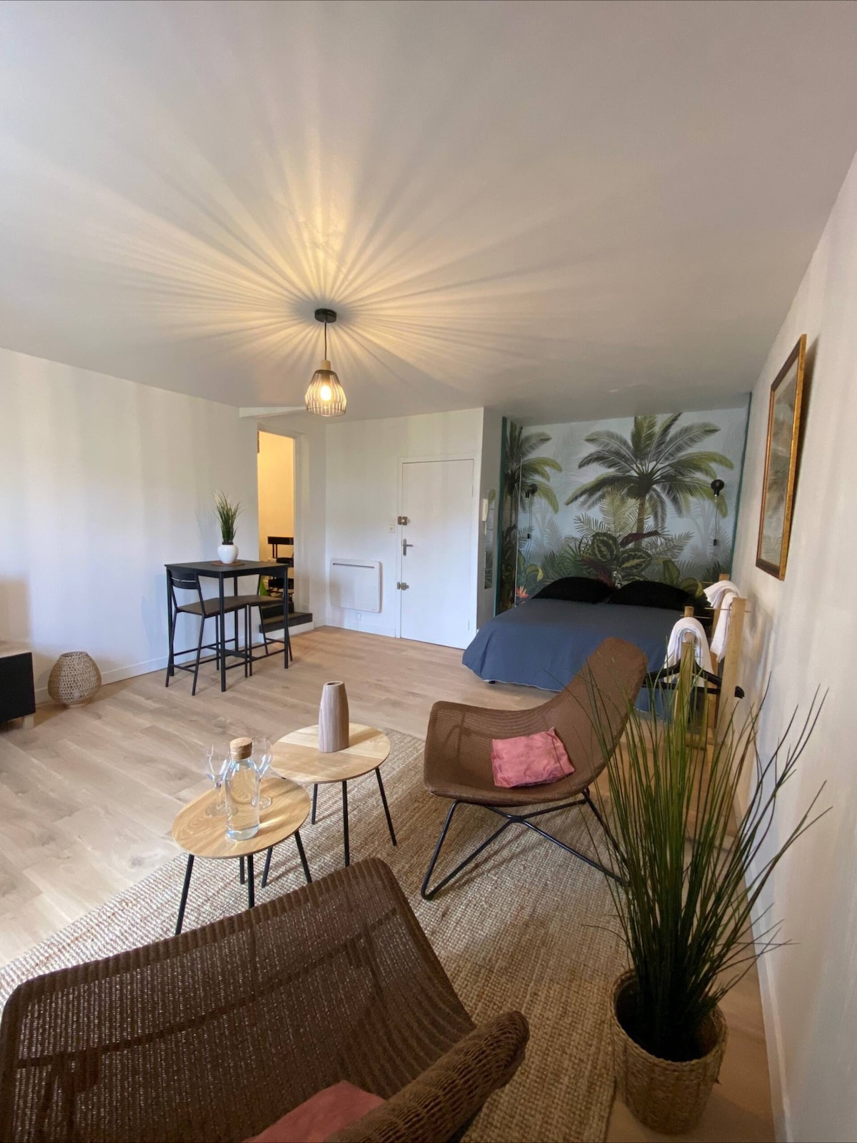 A spacious studio includes a designated seating area with two chairs and two small tables. A dining space is visible with a tall table and two stools. The bed is situated behind a partition, accented by a tropical mural, and soft lighting enhances the inviting atmosphere.