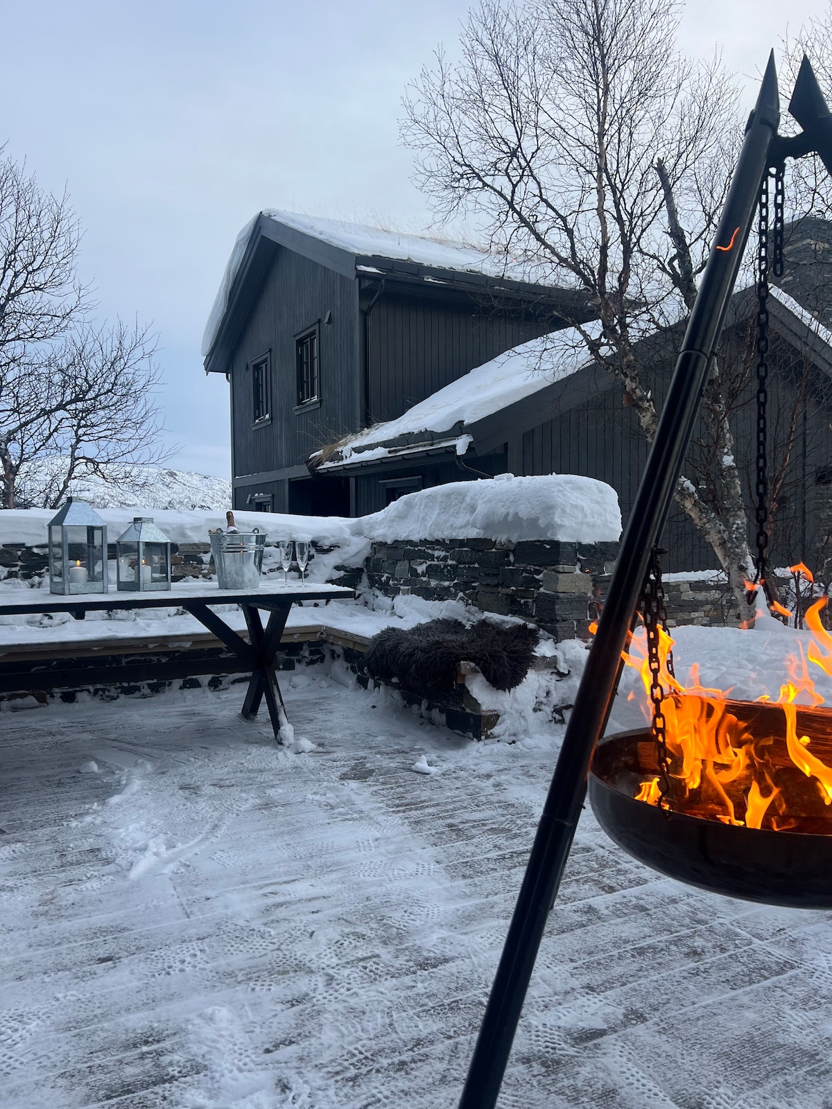 A snow-covered outdoor area features a firepit with flames, surrounded by a black metal structure. In the background, a dark wooden building can be seen, framed by trees without leaves. Lanterns are placed on a stone ledge, enhancing the serene winter scene.