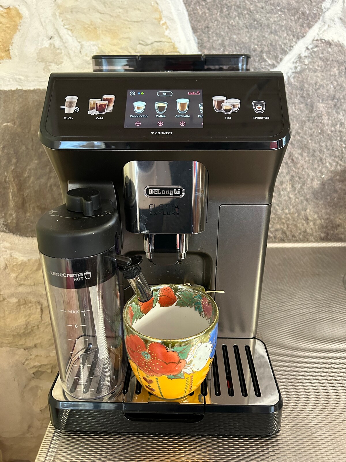 An espresso machine is positioned on a stainless steel countertop, featuring a touchscreen display that outlines various beverage options. A clear water container is attached, while a colorful mug, adorned with a strawberry design, is placed beneath the spout, ready for filling.