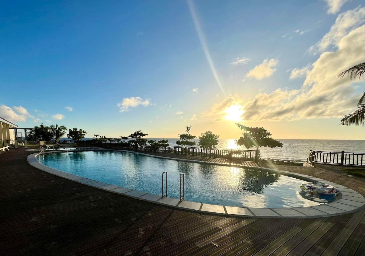 An expansive outdoor pool is situated near the ocean, reflecting the bright golden sunlight. Lush greenery and palm trees surround the area, enhancing the serene landscape. A wooden deck provides space for relaxation, contributing to the tranquil setting by the water.