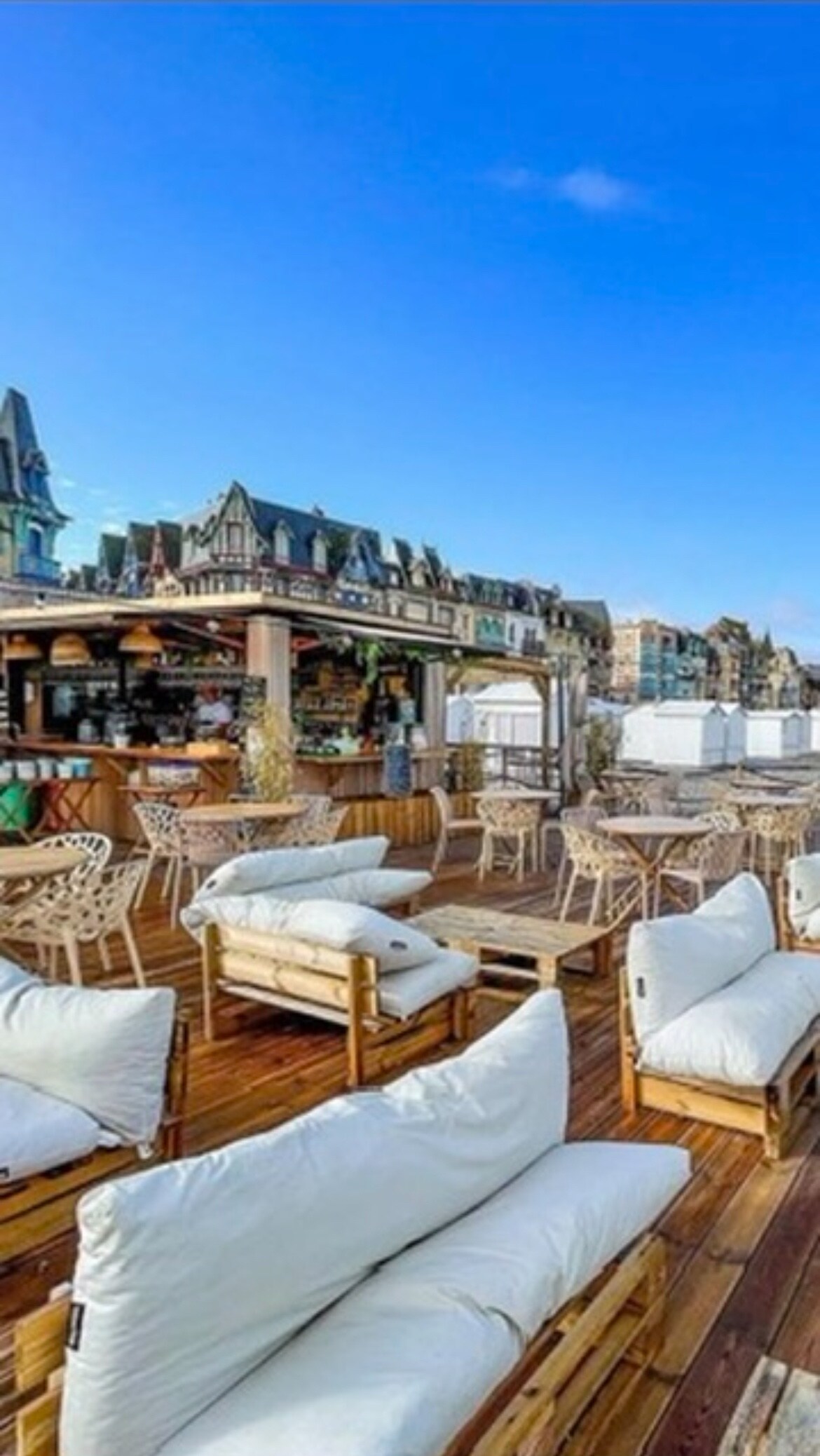 An outdoor seating area features comfortable lounge furniture made of natural wood. Soft white cushions are arranged around low tables, creating a relaxed atmosphere. The background showcases charming architectural details of neighboring buildings under a clear blue sky.
