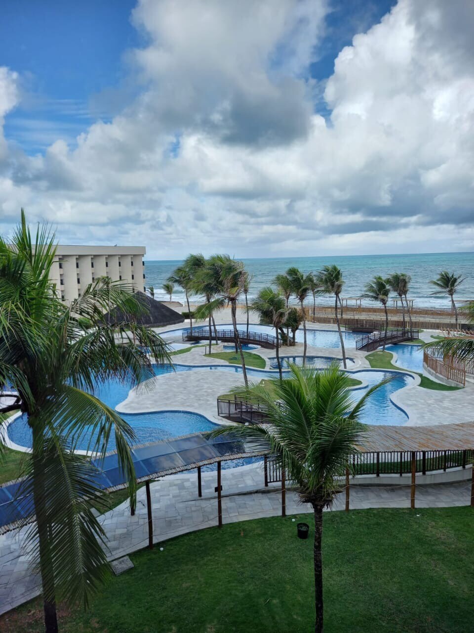 The image captures a resort pool area surrounded by palm trees, leading to the beach. Multiple pools of varying shapes and sizes are visible, with clear blue water. The ocean and a cloudy sky are presented in the background, emphasizing a serene coastal environment.