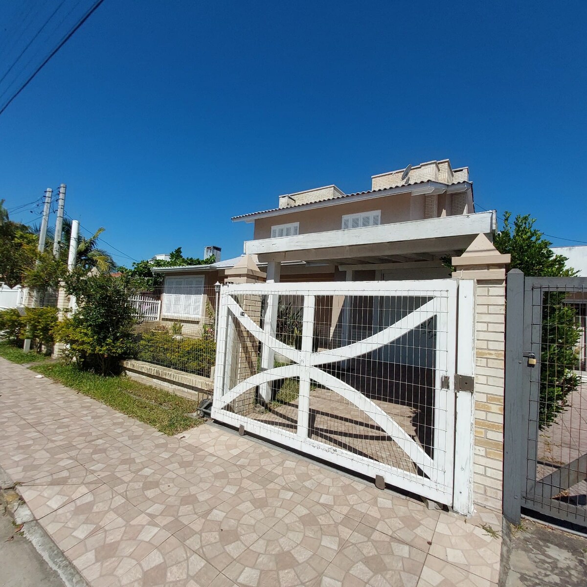 A two-story house is framed by a white fence, with a gated entrance visible. The façade features large windows and a light-colored exterior, surrounded by lush greenery and well-maintained landscaping. A clear blue sky provides a bright backdrop.